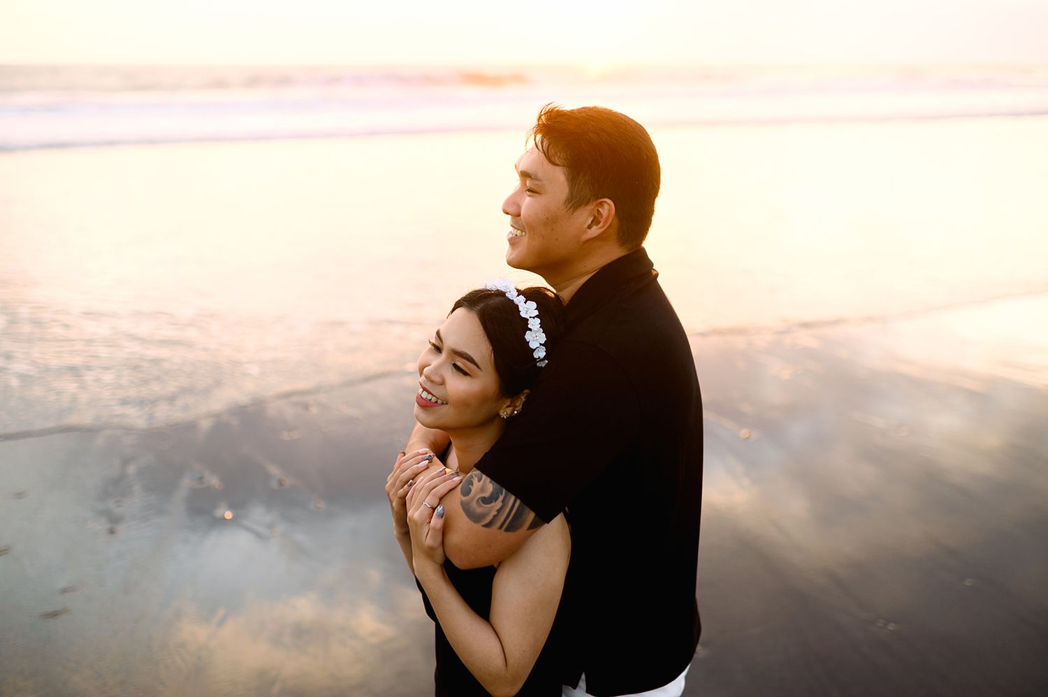 Couple embracing romantically on a beach at sunset, woman smiling with eyes closed, man in black shirt.