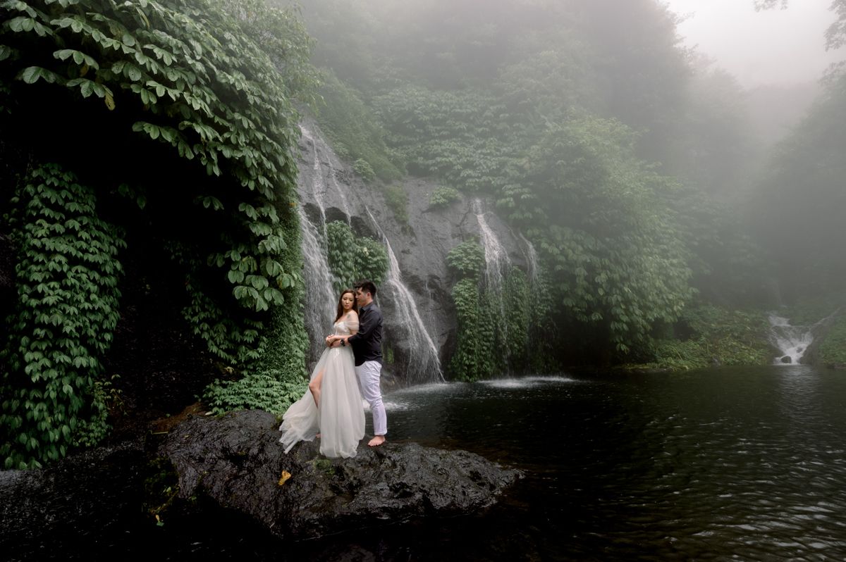 Couple embracing on misty rocks beside a lush waterfall surrounded by green ivy in a romantic wedding photo.