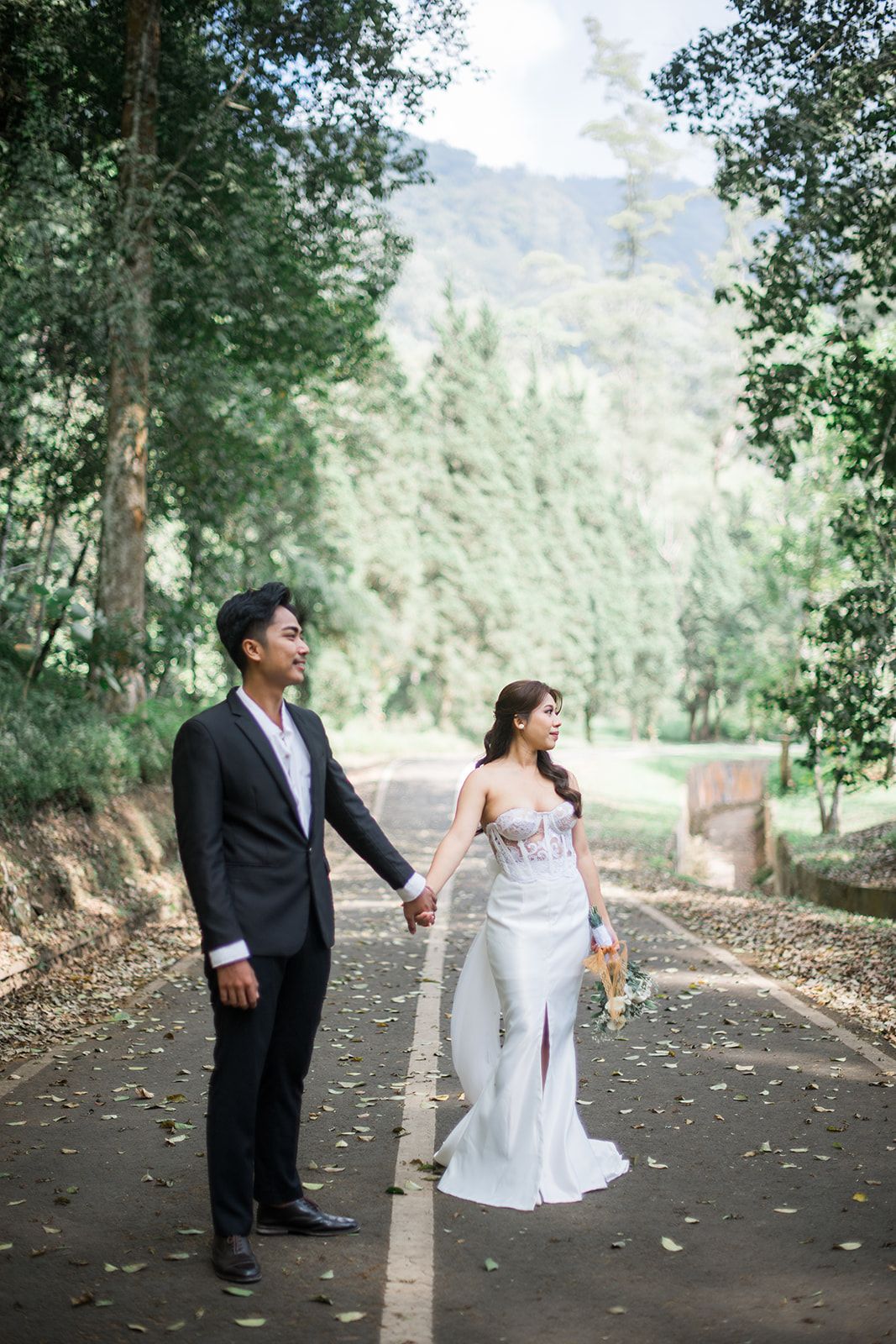 A bride and groom hold hands on a tree-lined path, with mountains in the background, during an outdoor wedding photoshoot.