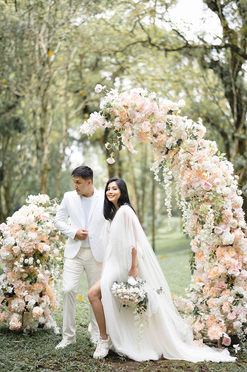 Couple in white wedding attire poses under a floral arch with pink blooms in a lush forest setting.