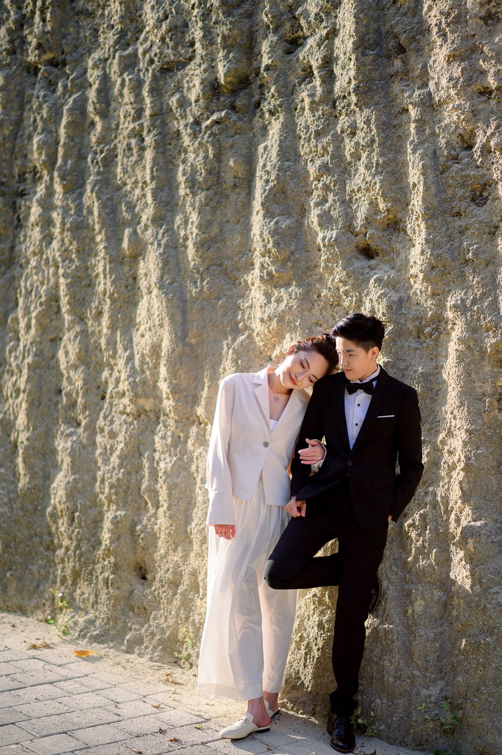 A couple in wedding attire poses against a textured stone wall, the bride in white suit and groom in black tuxedo.
