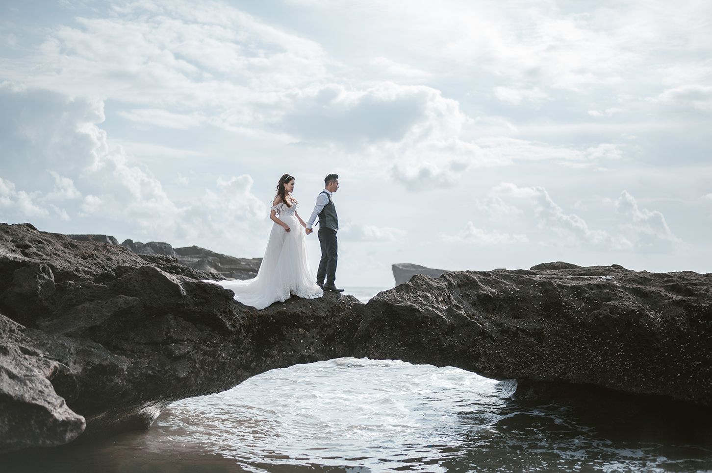 Bride and groom holding hands on rocky coastal arch with ocean waves and dramatic cloudy sky backdrop.