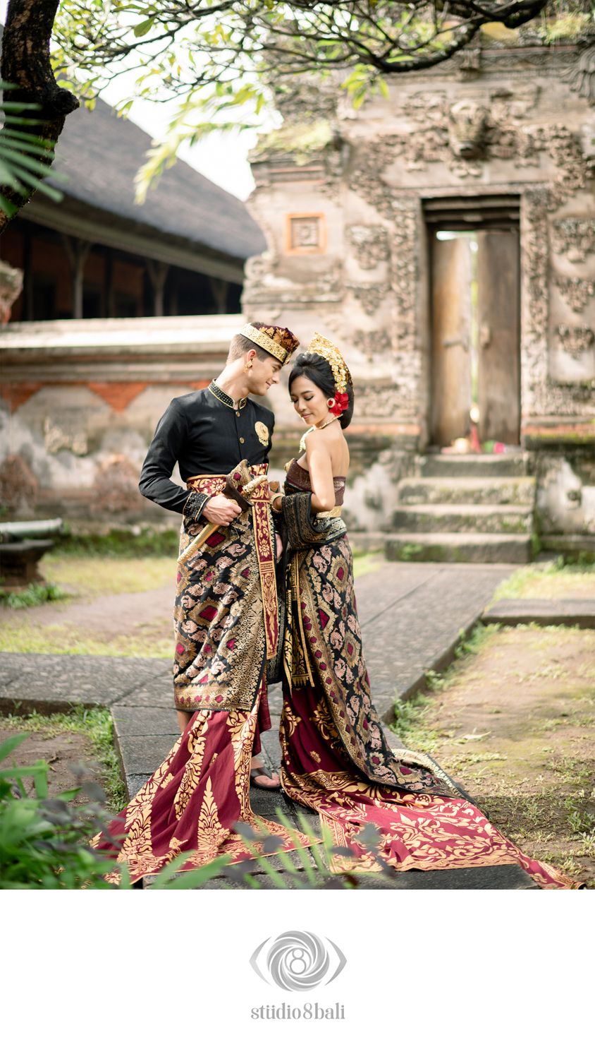Couple in traditional Balinese wedding attire posing at an ancient temple, wearing ornate batik garments and gold accessories.