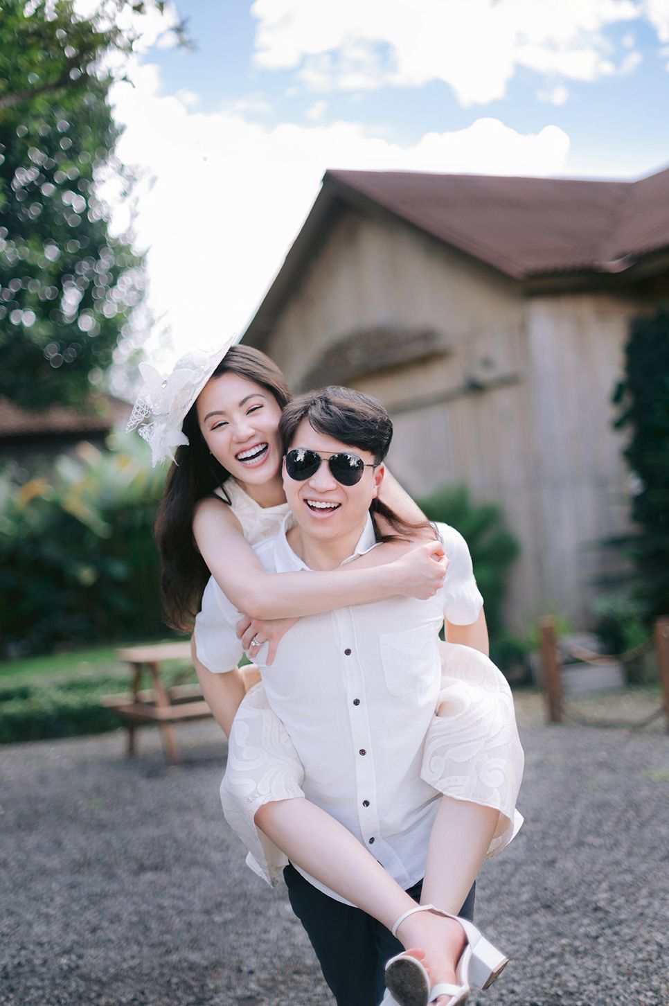 Happy couple laughing outdoors, man giving woman a piggyback ride, both wearing white outfits and sunglasses.