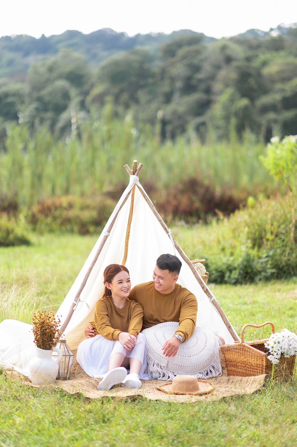 Couple sitting together in a boho teepee tent on a picnic blanket in a lush green meadow.