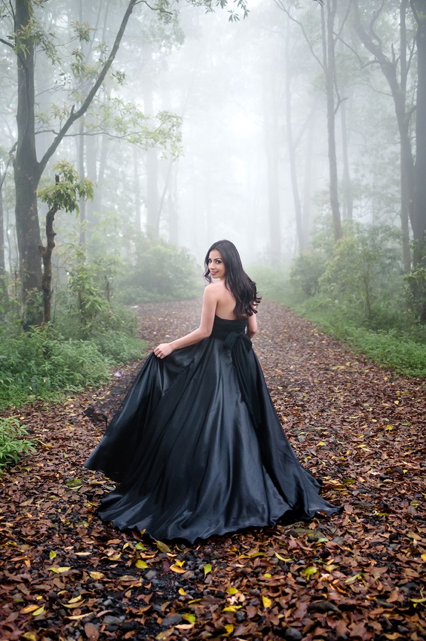 Woman in black ball gown standing on a foggy forest path covered in autumn leaves, glancing back over her shoulder.