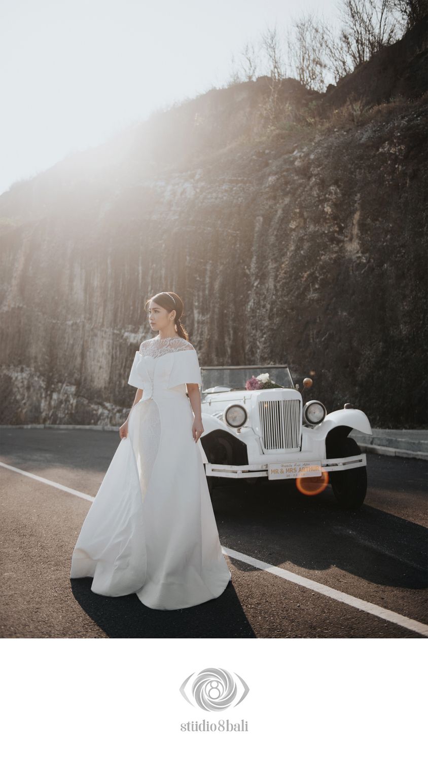 Bride in elegant white gown poses on road beside vintage white car with rocky cliffs and sunlight in background.
