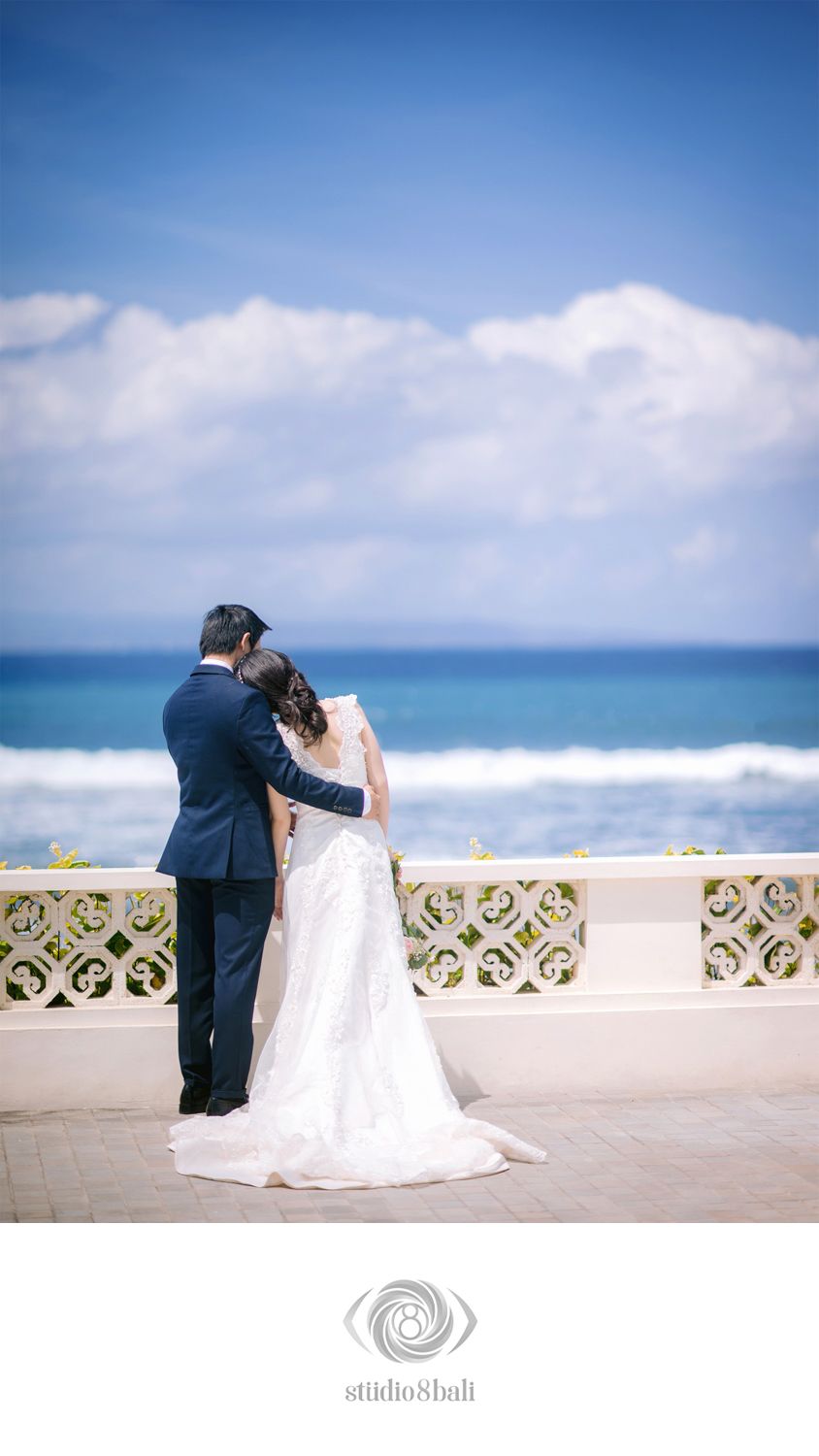 Bride and groom stand together on ocean-view terrace, bride in white gown with veil, groom in navy suit, Bali wedding.
