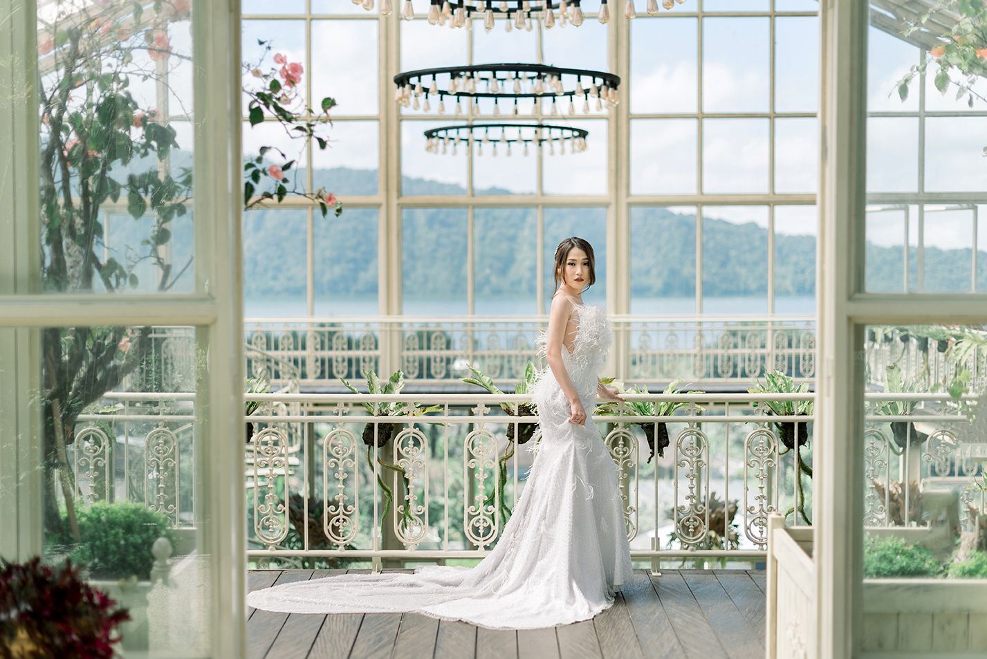 Elegant bride in white gown standing on ornate balcony with chandelier and scenic water view through large windows.