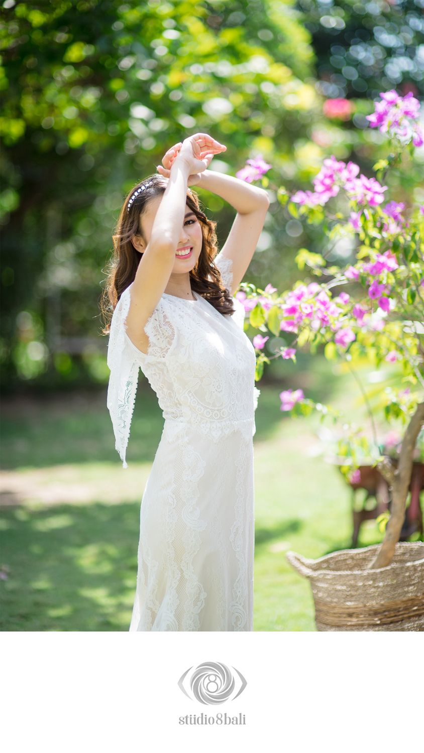 Bride in white lace dress posing among pink flowers in a lush garden, shielding her eyes from sunlight.