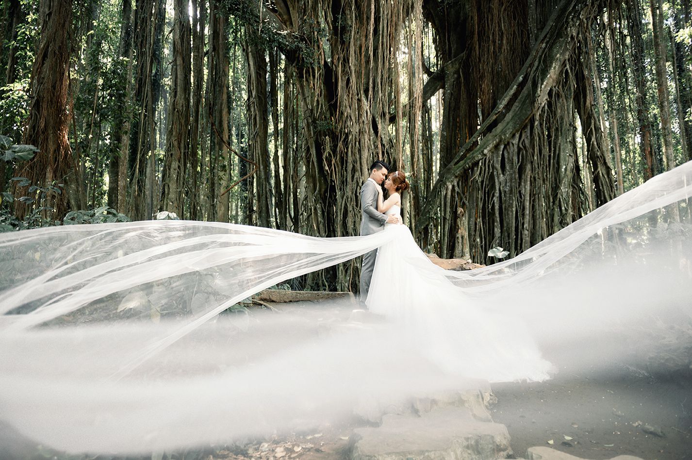 A couple shares a romantic kiss in a forest as the bride's dramatic white veil billows in the wind.