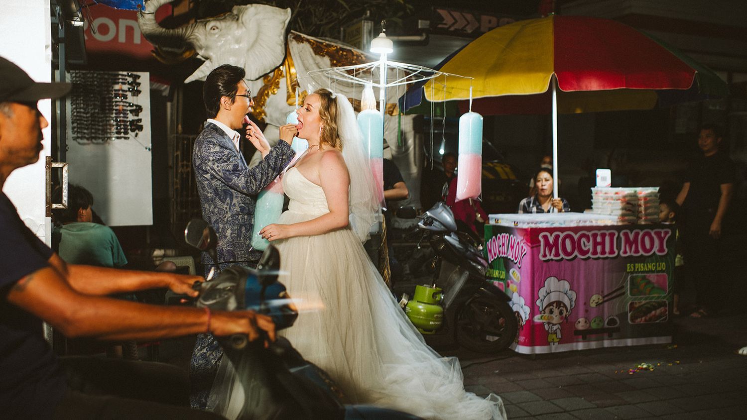 Bride and groom share a romantic moment at a colorful night market street food stall.