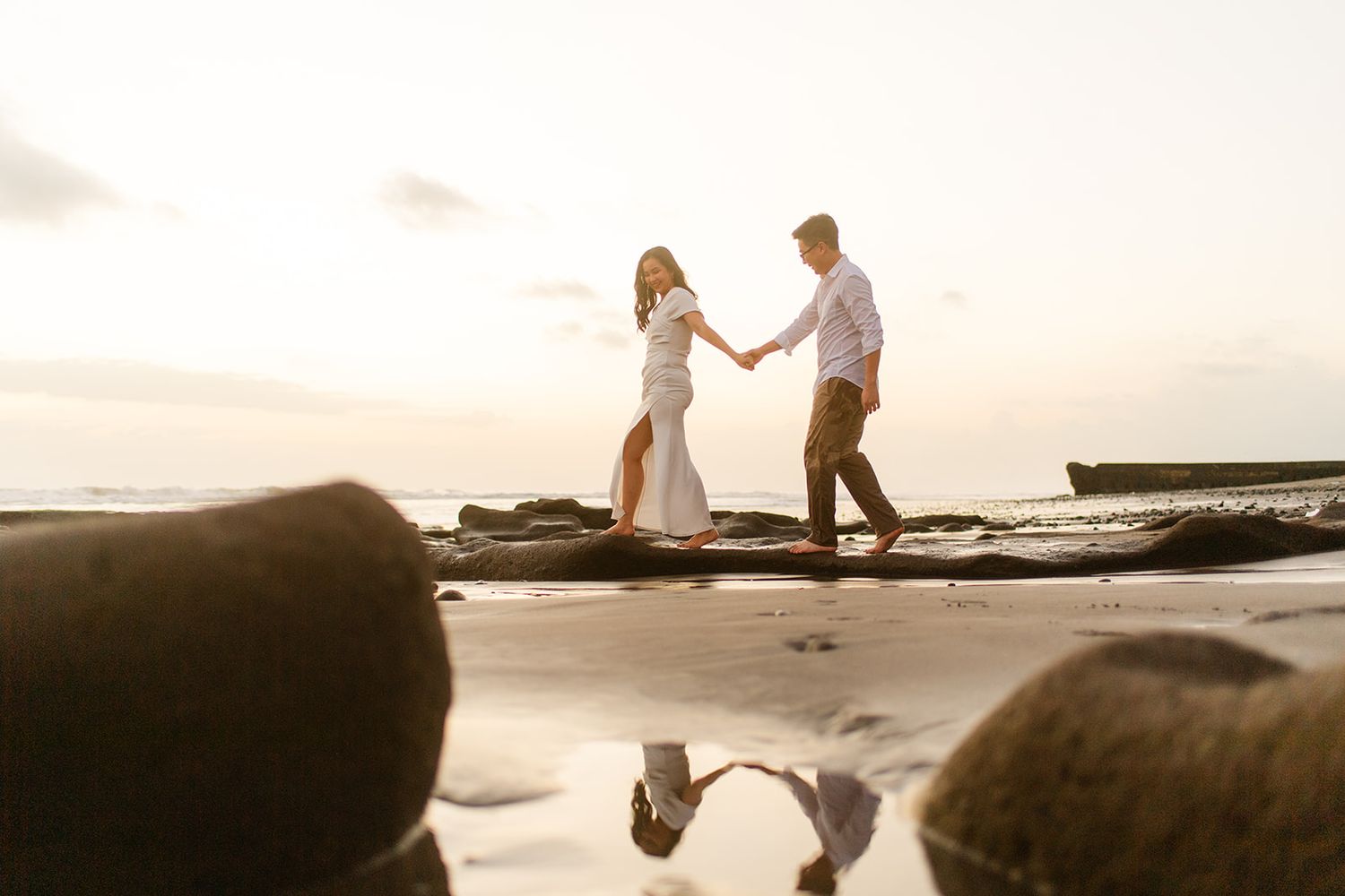 Couple holding hands on beach rocks at sunset with reflection in water.
