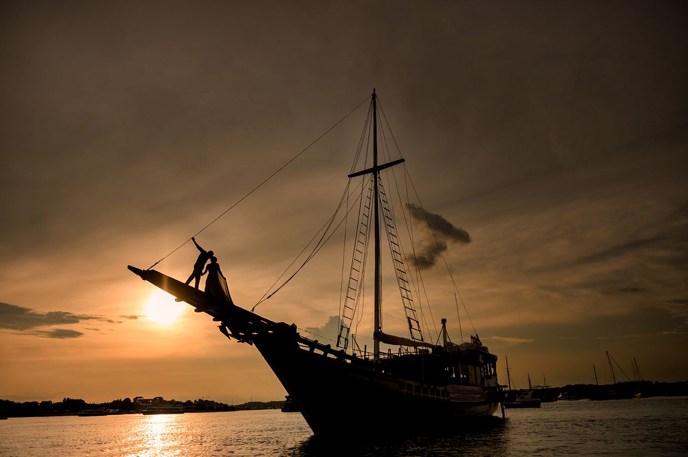Silhouette of a tall sailing ship anchored at sea during a dramatic golden sunset with cloudy skies.