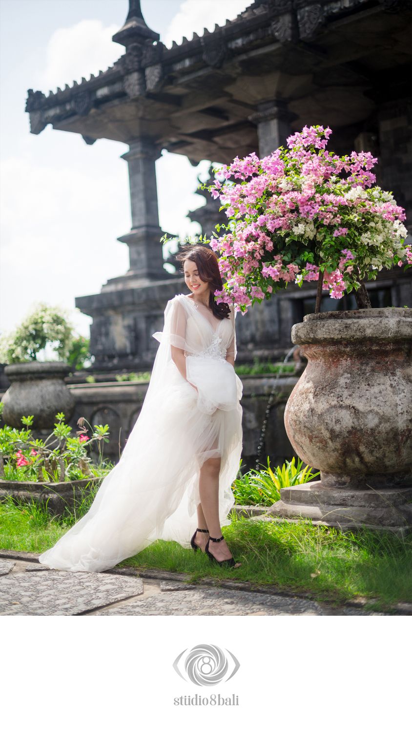 Bride in white wedding gown poses by a large stone urn with pink flowers at a Bali temple garden.