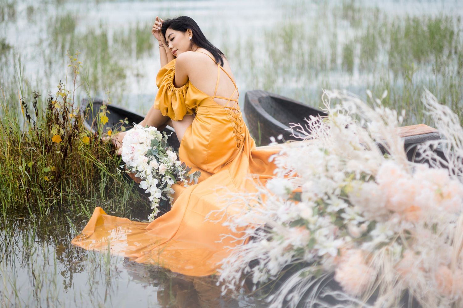 Woman in orange gown sits in a boat surrounded by wildflowers in a serene water landscape.