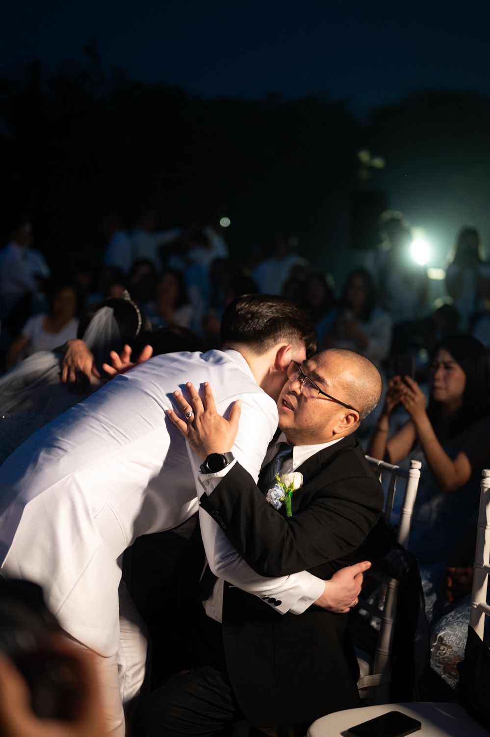 Groom in white suit embraces older man in black tuxedo with boutonniere at outdoor evening wedding ceremony.