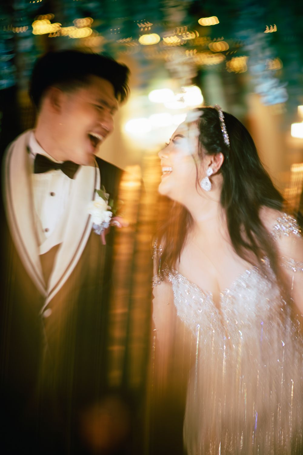 A joyful bride and groom share a laughing moment, she in a beaded gown, he in a tuxedo, surrounded by warm bokeh lights.