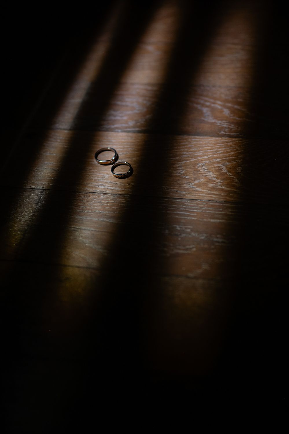 Two wedding rings resting on a wooden floor, dramatically lit by streaks of light and shadow.