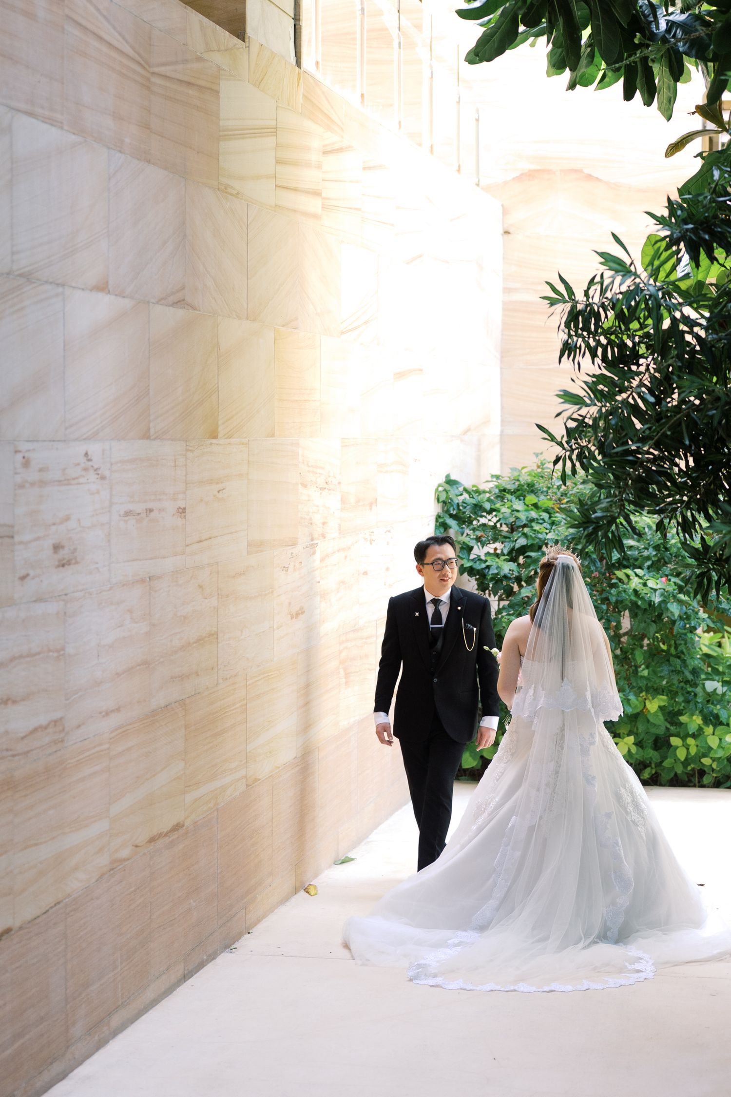 A bride in a white gown and veil walks with a groom in a black suit along a marble wall with lush greenery.
