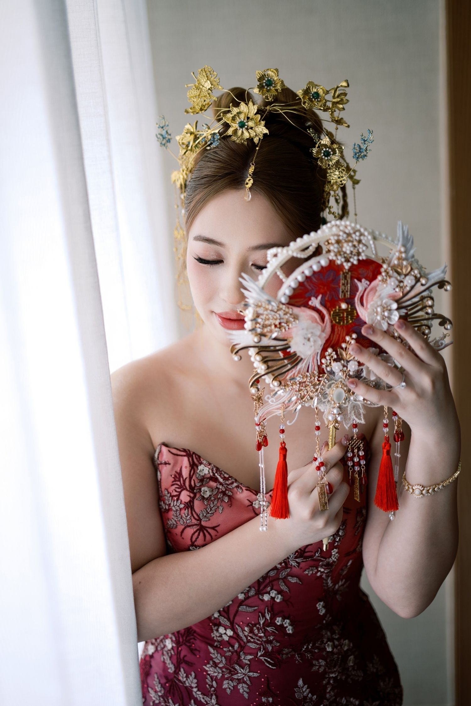 A bride in a red floral qipao holds a decorative fan, wearing a gold headdress in traditional Chinese bridal attire.