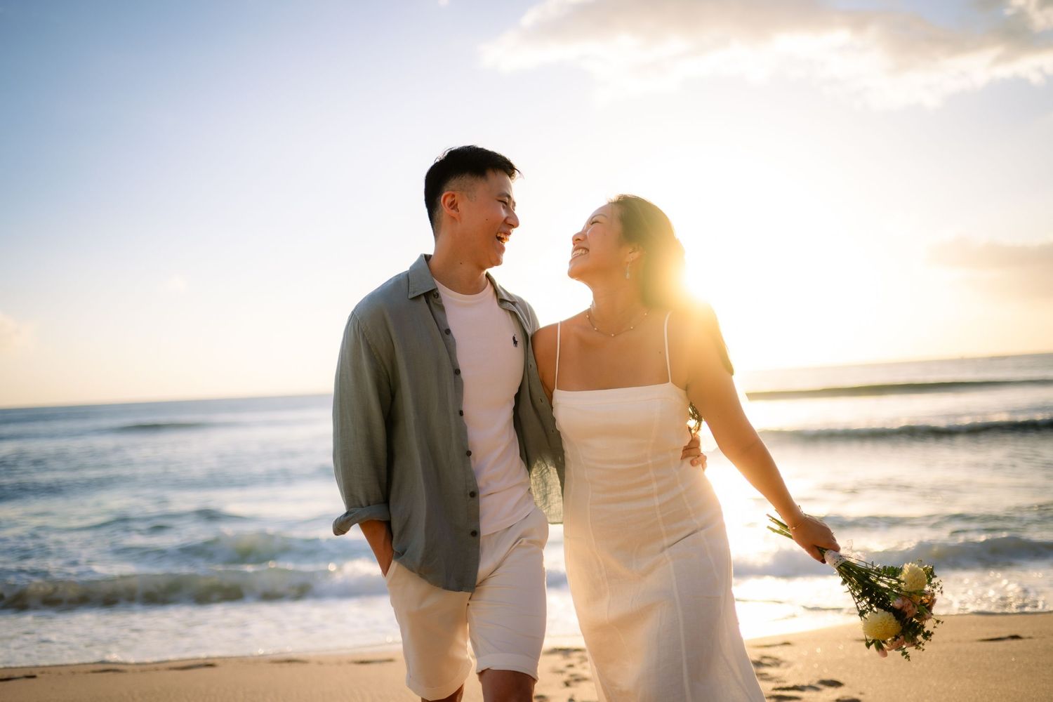 Couple walking on a beach at sunset, woman in white dress holding flowers, man in gray shirt, ocean waves in background.
