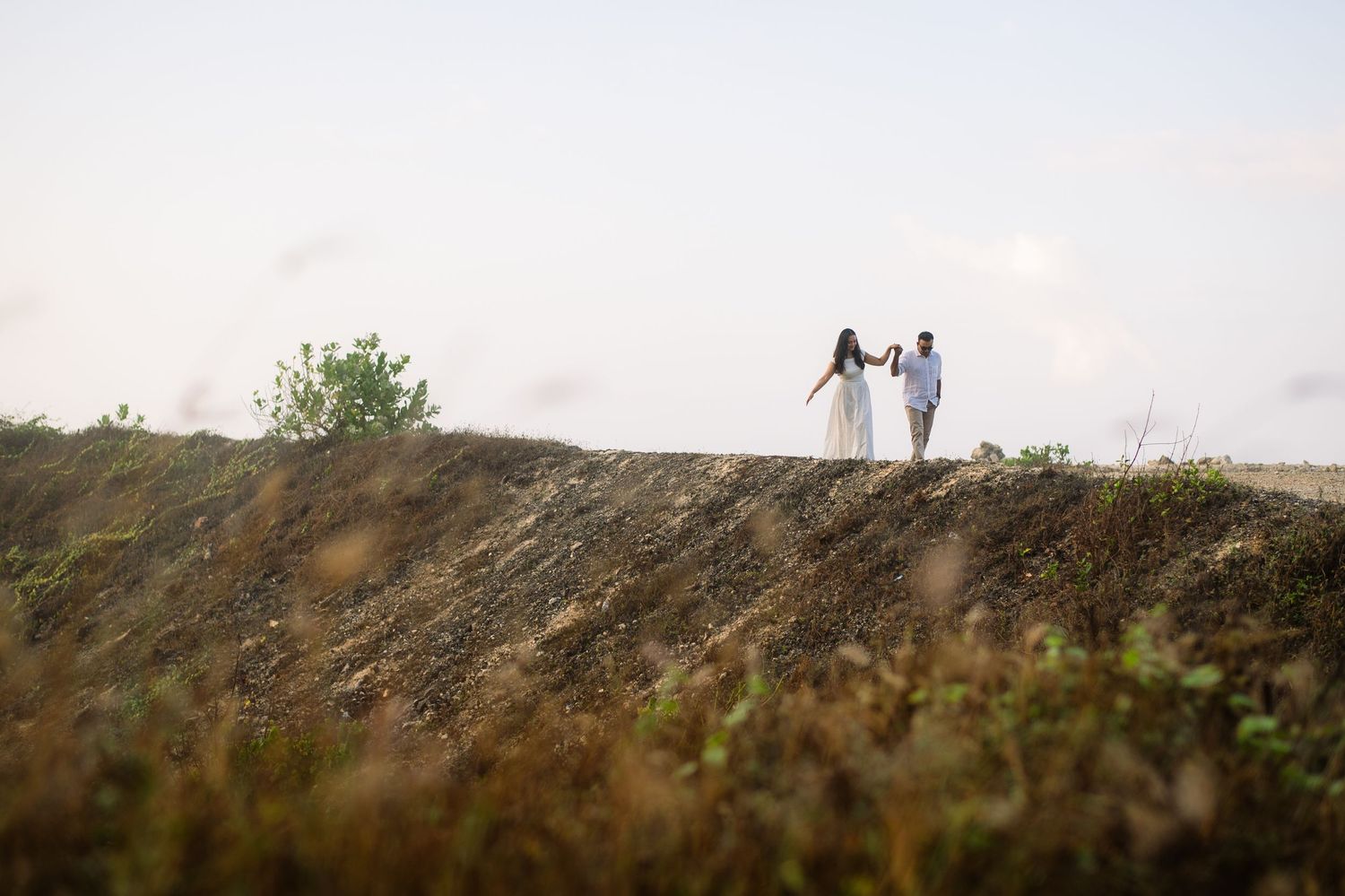 Two women in white dresses walk hand in hand along a grassy hillside under a soft sky.