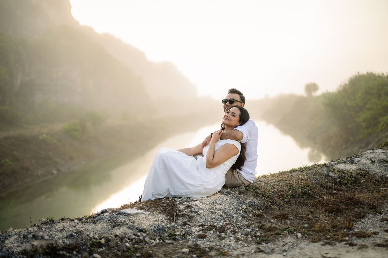 Romantic couple in white embracing on rocky cliff with misty river and green hills behind them.