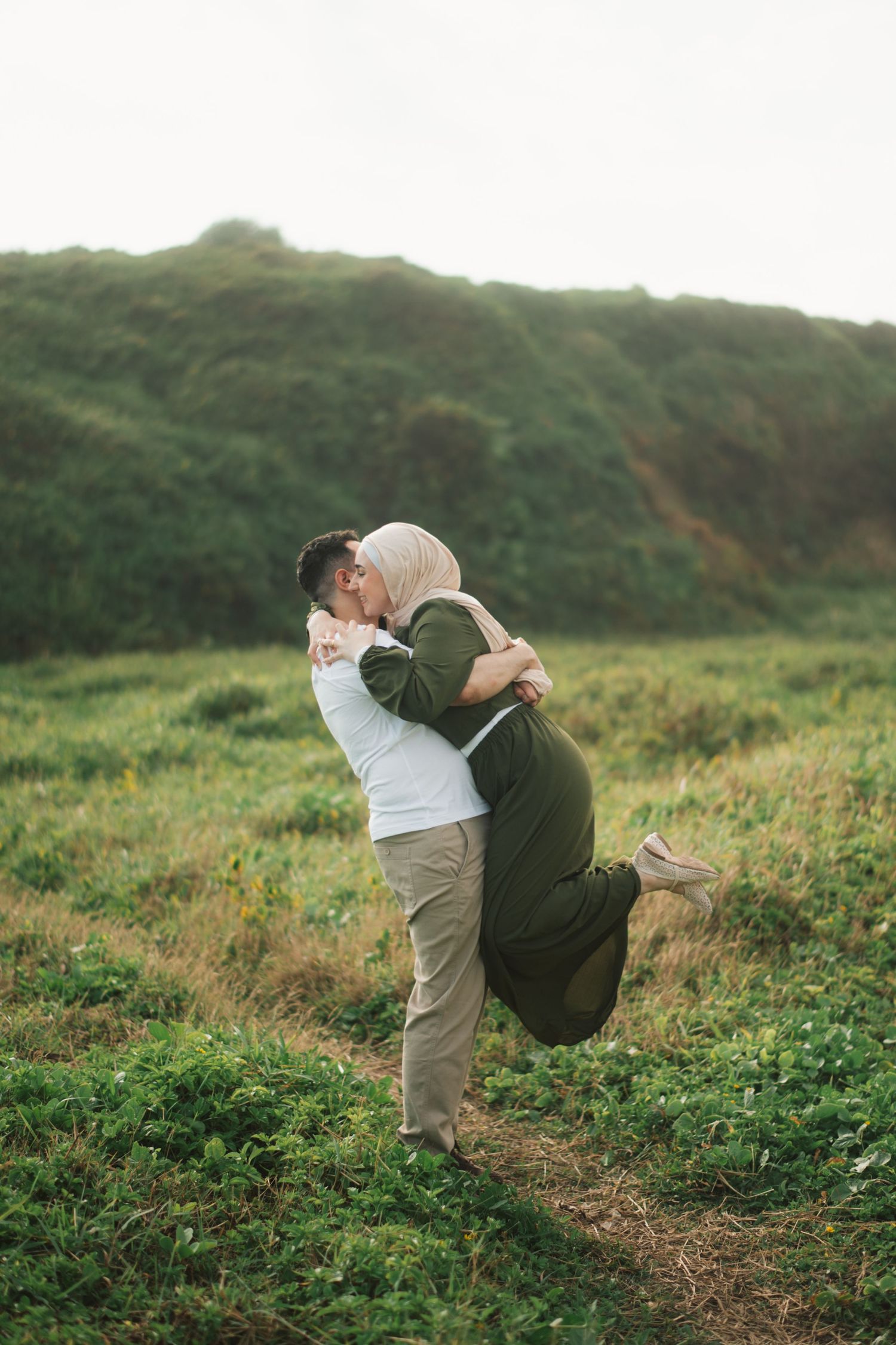 A couple sharing a romantic kiss in a lush green field, man lifting woman wearing hijab and olive dress.