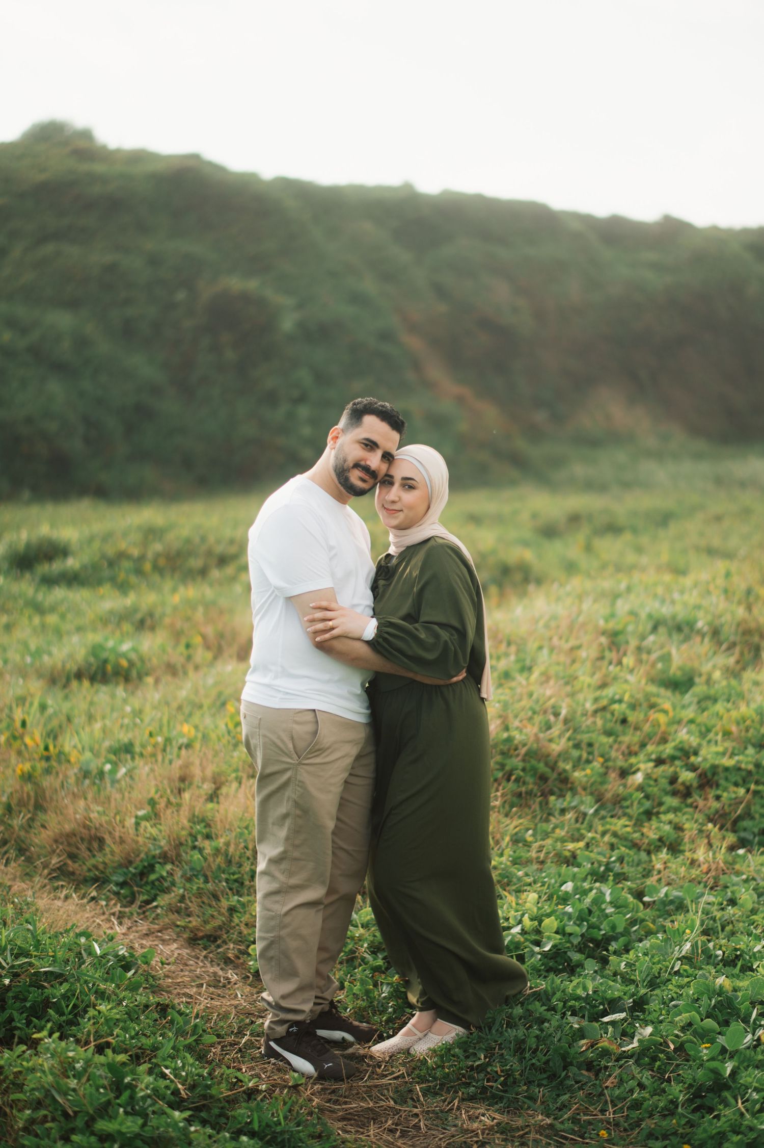 A couple embracing in a green field, woman in hijab and olive dress, man in white shirt, with hills behind.