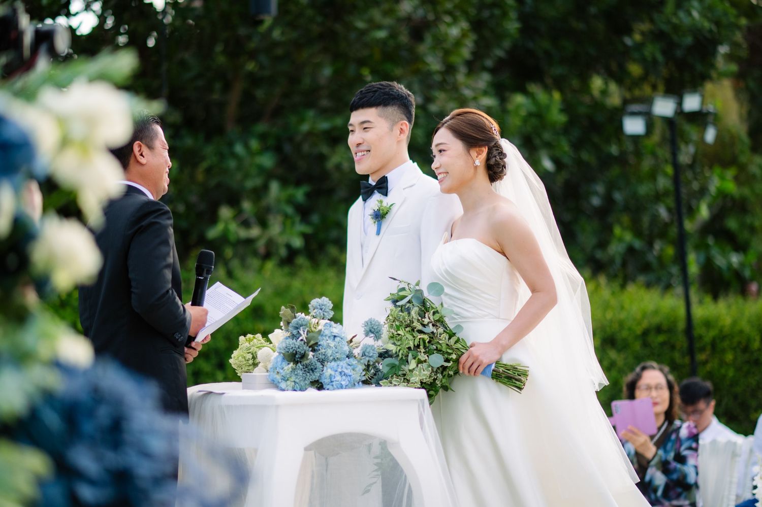 Bride and groom smile during an outdoor garden wedding ceremony while an officiant reads vows and guests watch nearby.