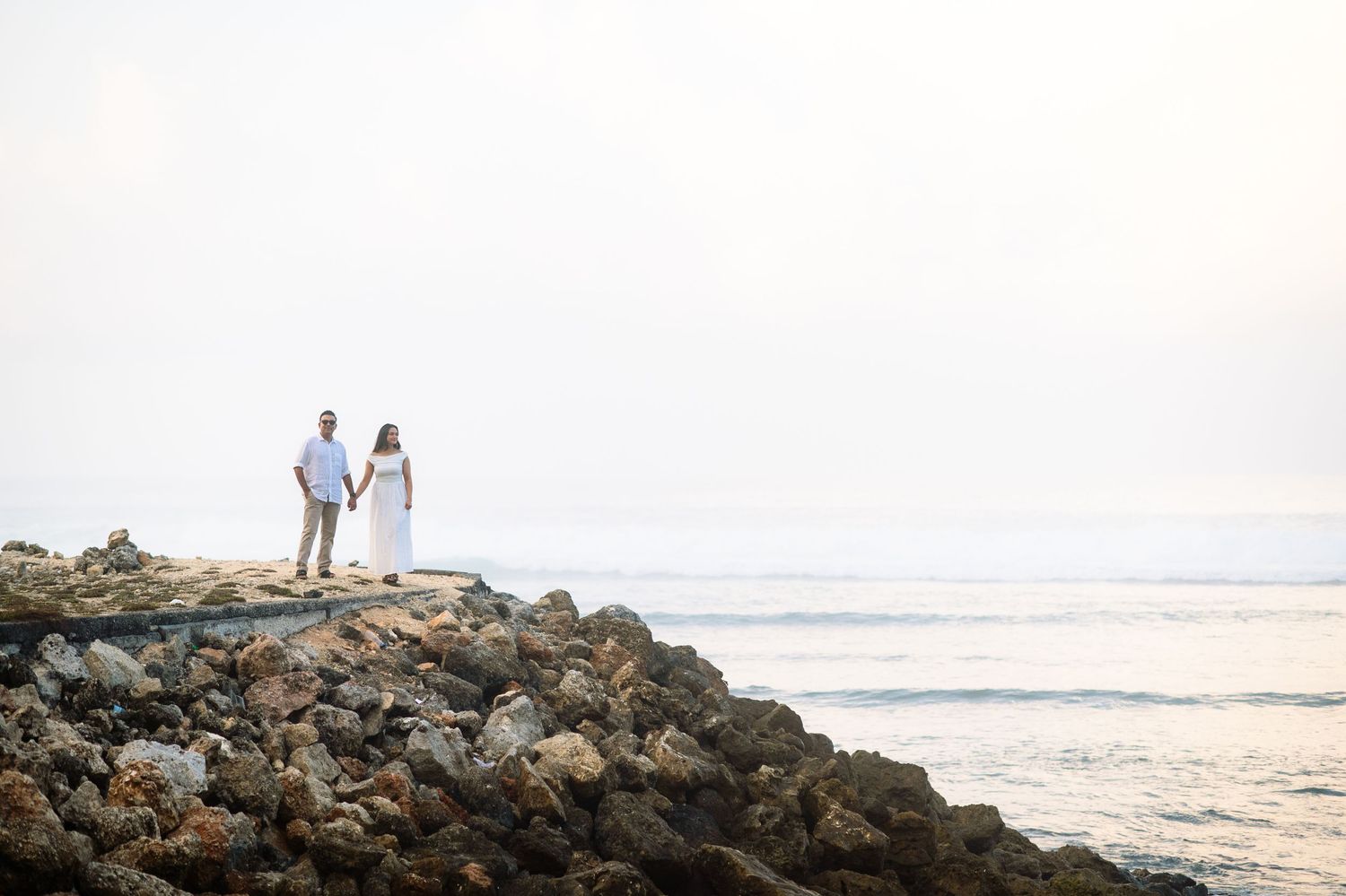 Couple in white attire standing on rocky shoreline at sunset with ocean waves in the background.