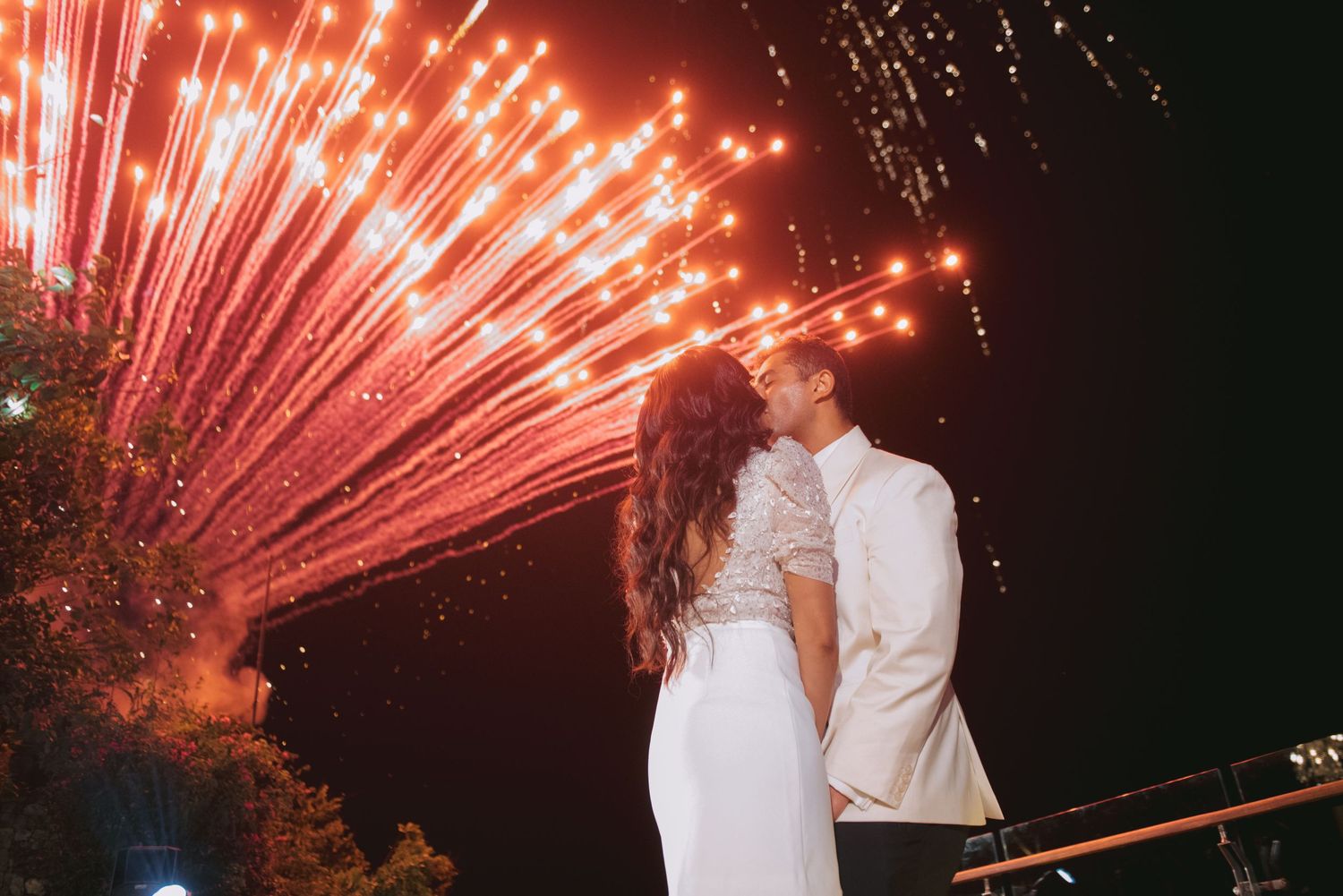 Romantic couple sharing a kiss with vibrant red fireworks lighting the night sky behind them.