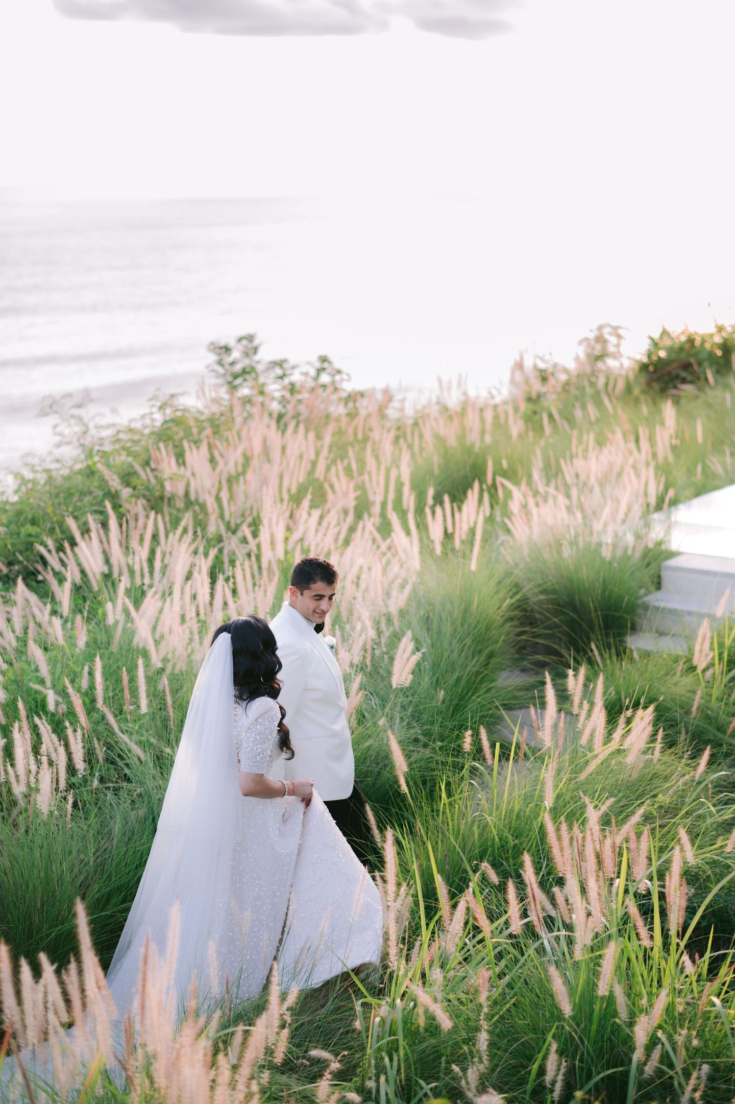 A bride and groom share a romantic moment among tall ornamental grasses with ocean views at their outdoor wedding.