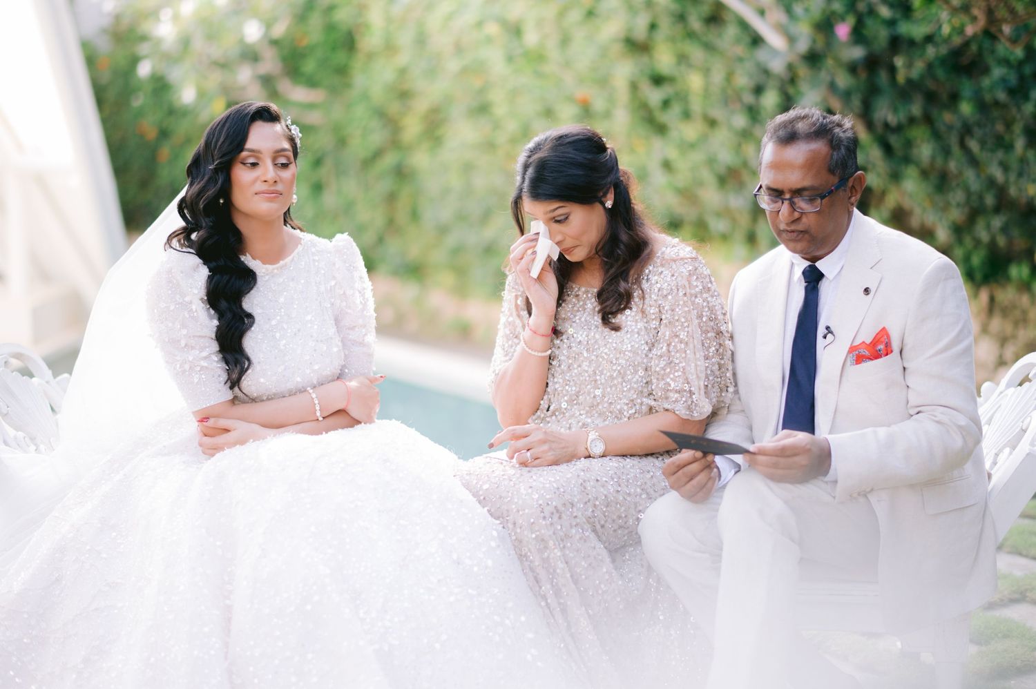 Two brides in white gowns sit with emotional family member at outdoor wedding ceremony.