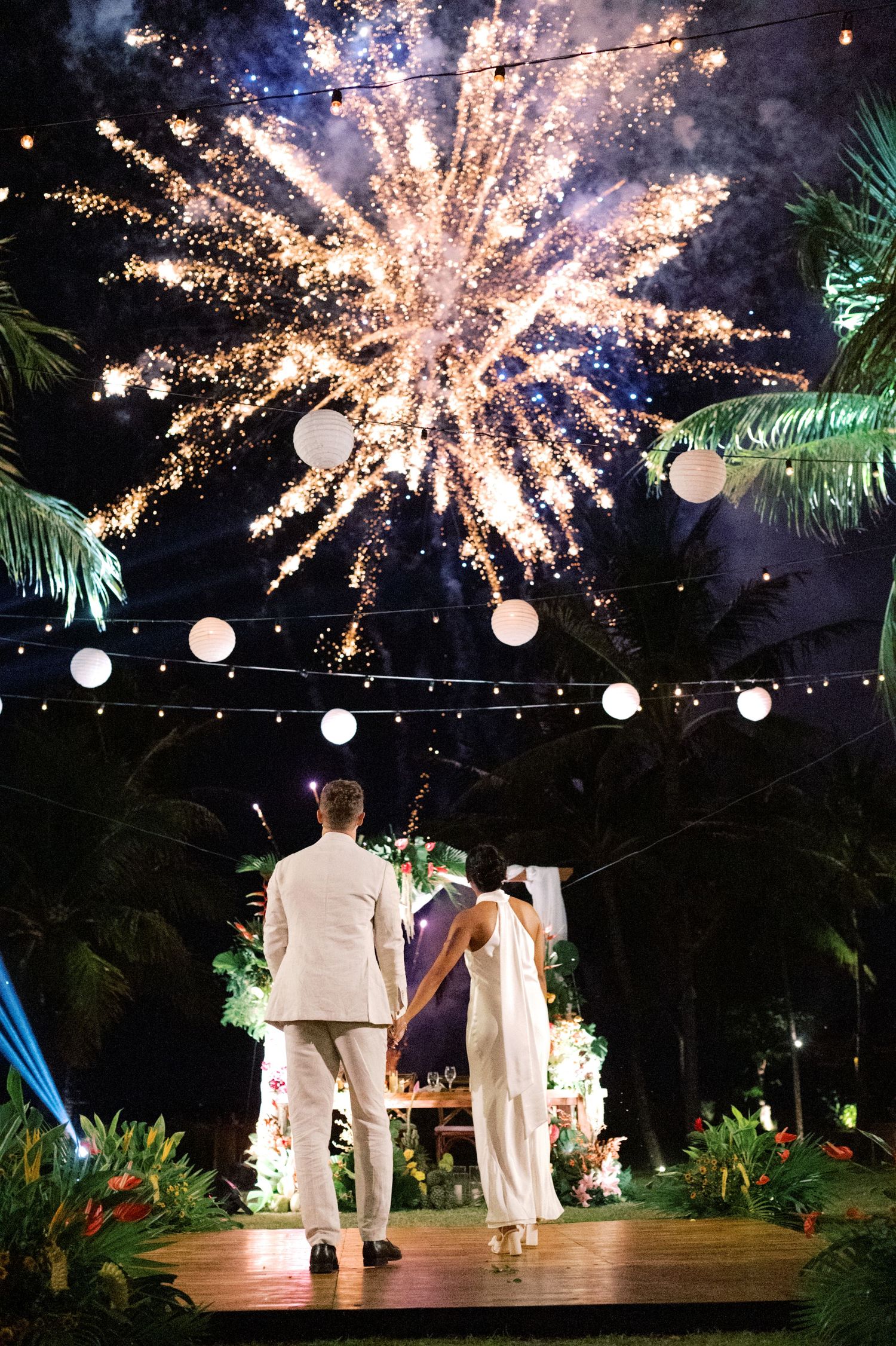 Bride and groom watch spectacular fireworks display at tropical outdoor wedding reception at night.