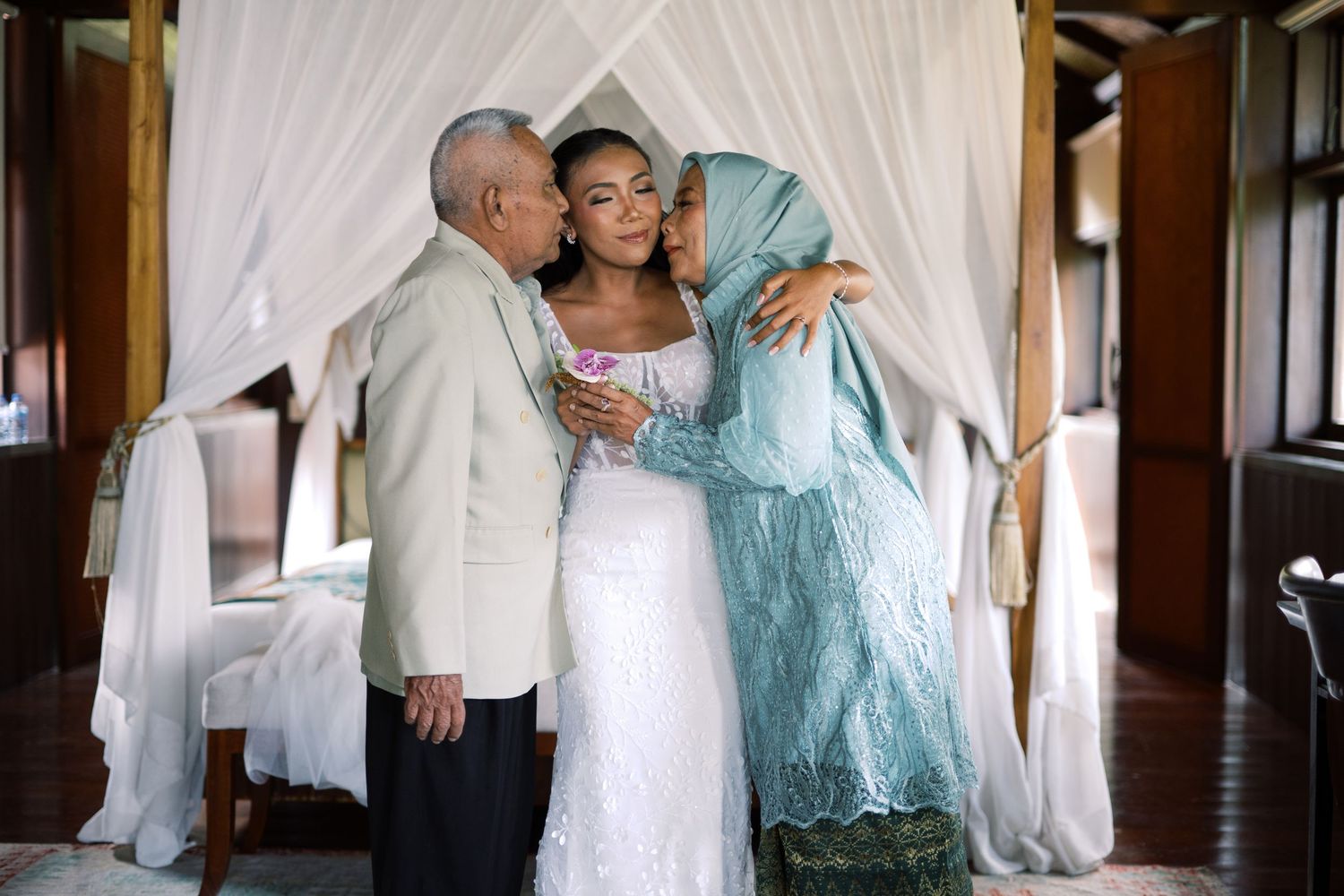 Bride in white lace gown receives kisses from parents under canopy bed in tropical villa.