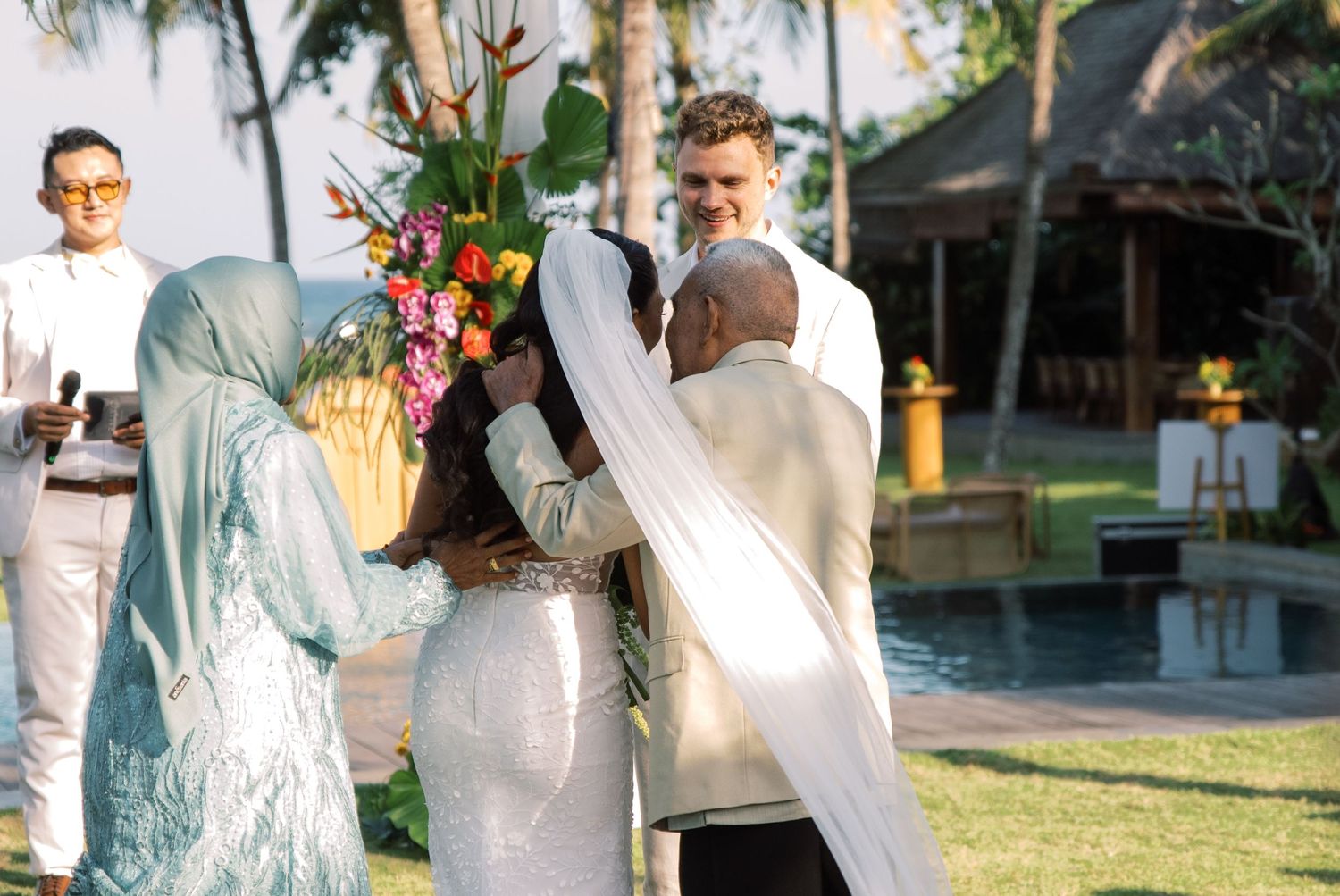 Bride receives blessing from family members during outdoor tropical wedding ceremony by pool.