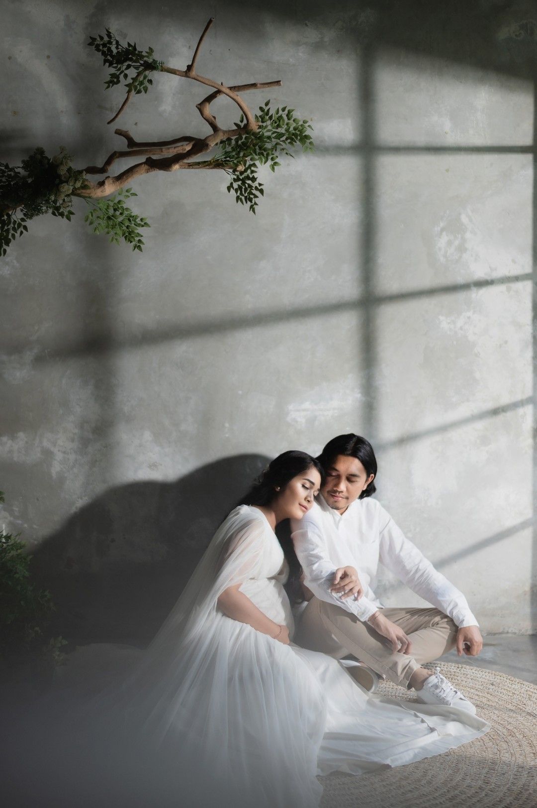 A couple in white attire sits together on a woven mat, surrounded by soft light and greenery in an elegant studio setting.