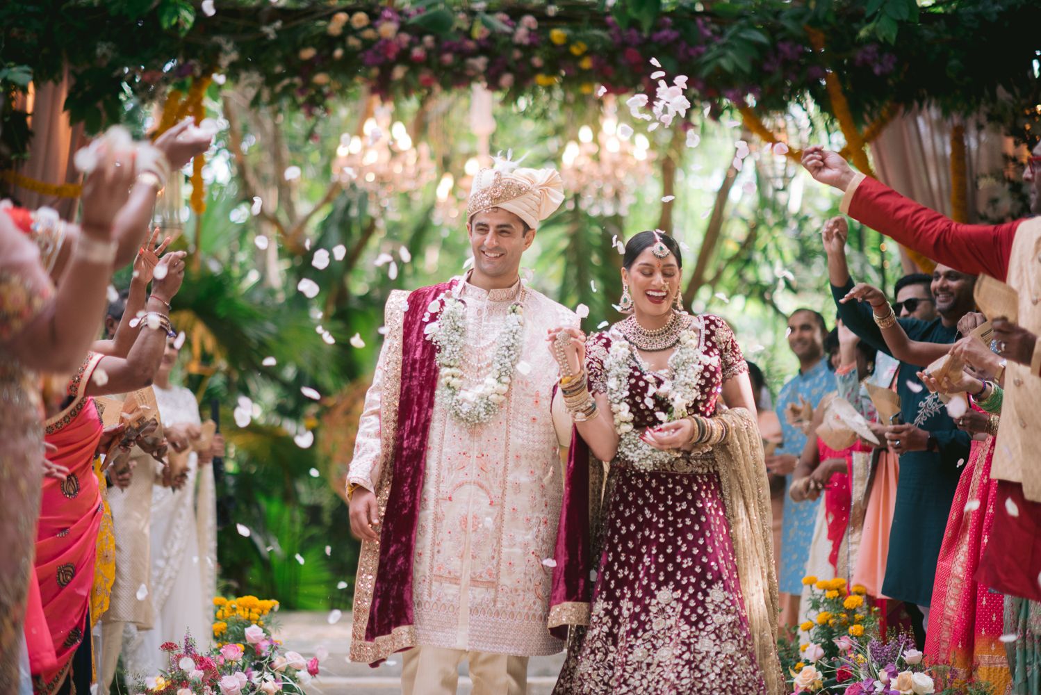 Couple walks through floral arch at Indian wedding ceremony, groom in white sherwani, bride in maroon lehenga.
