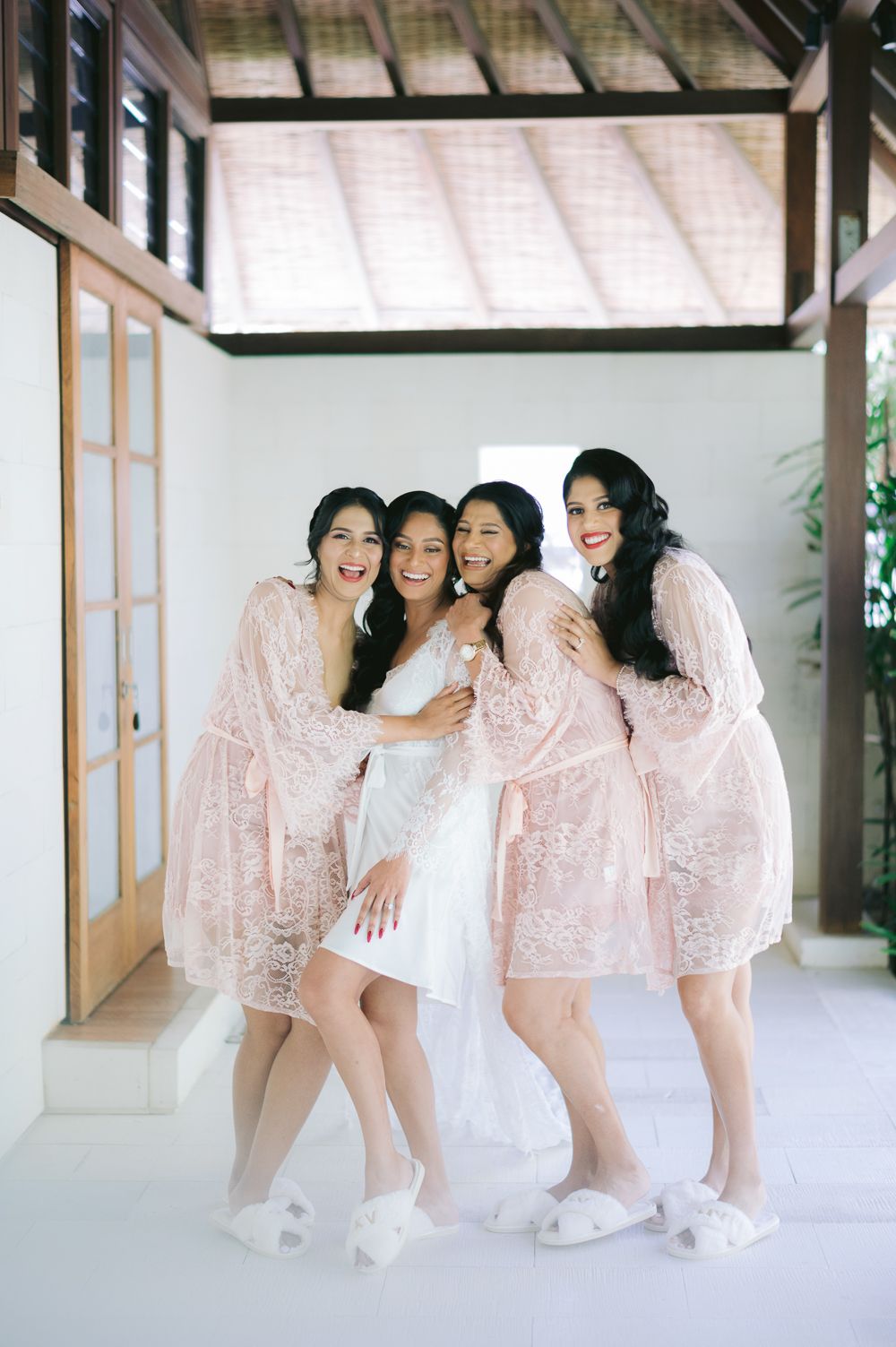 Bride in white dress with three bridesmaids in pink lace robes and fluffy slippers posing together indoors.