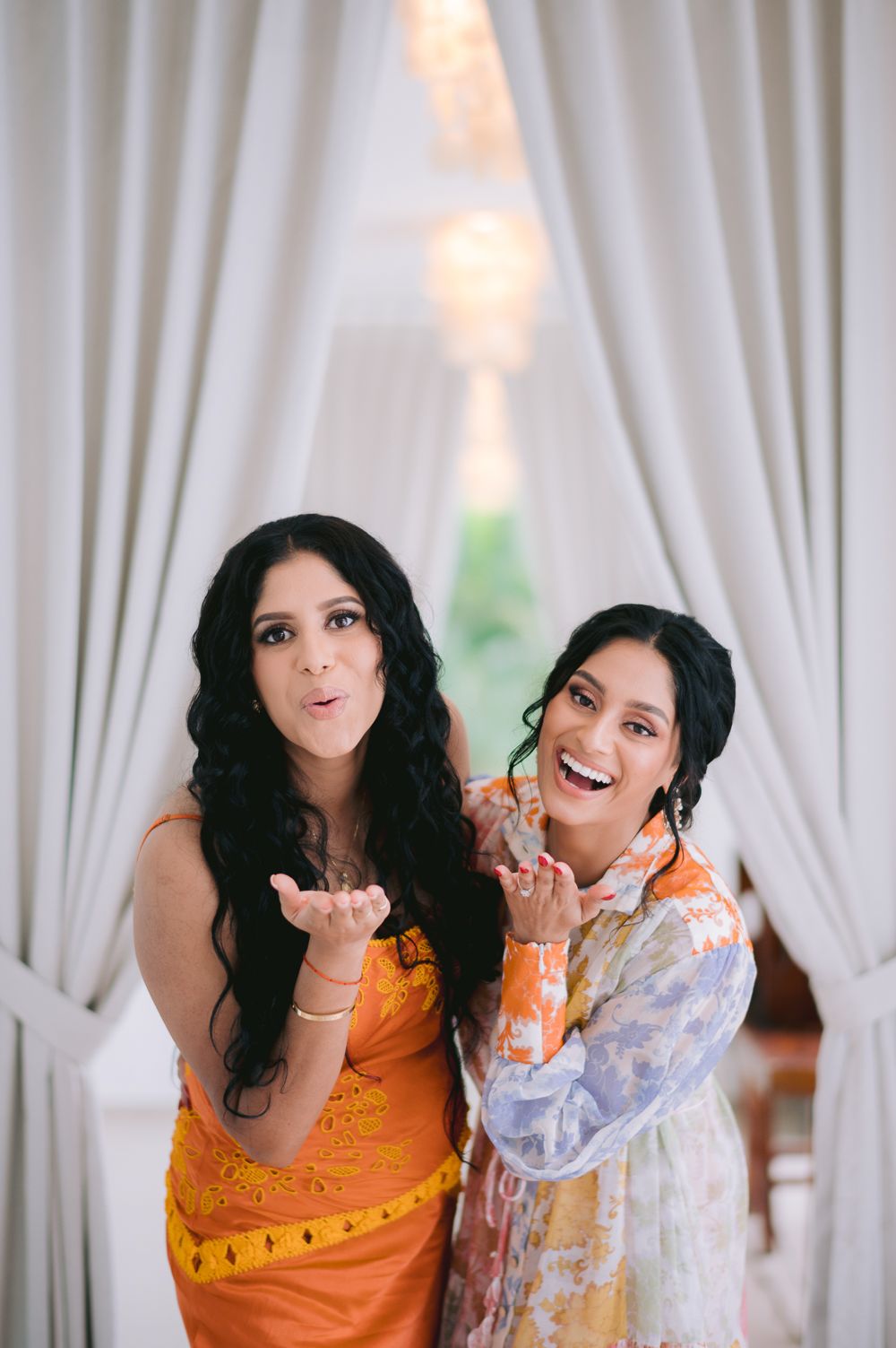 Two women in colorful dresses blowing kisses, posing joyfully indoors with white curtains backdrop.