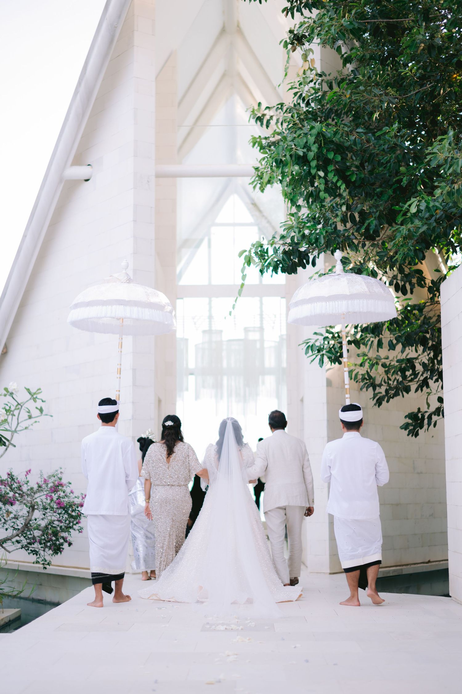 Bride in white gown and veil walks toward chapel entrance with wedding party carrying white ceremonial umbrellas.