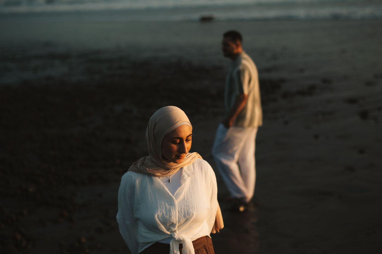 A woman in a white hijab and man stand on a beach at golden hour in flowing white garments.