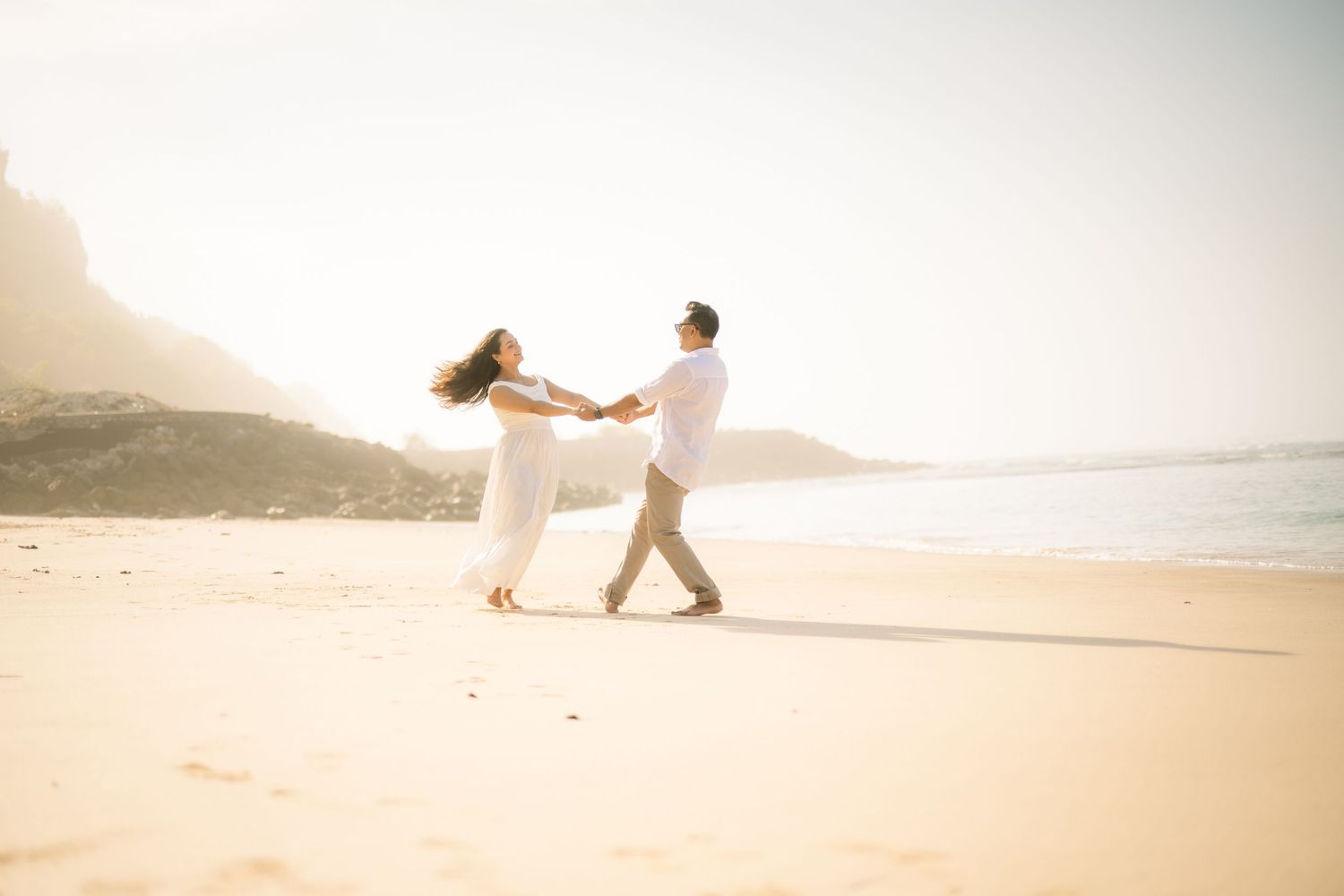 Couple dancing joyfully on a sunny beach, woman in white dress, golden sand and ocean backdrop.