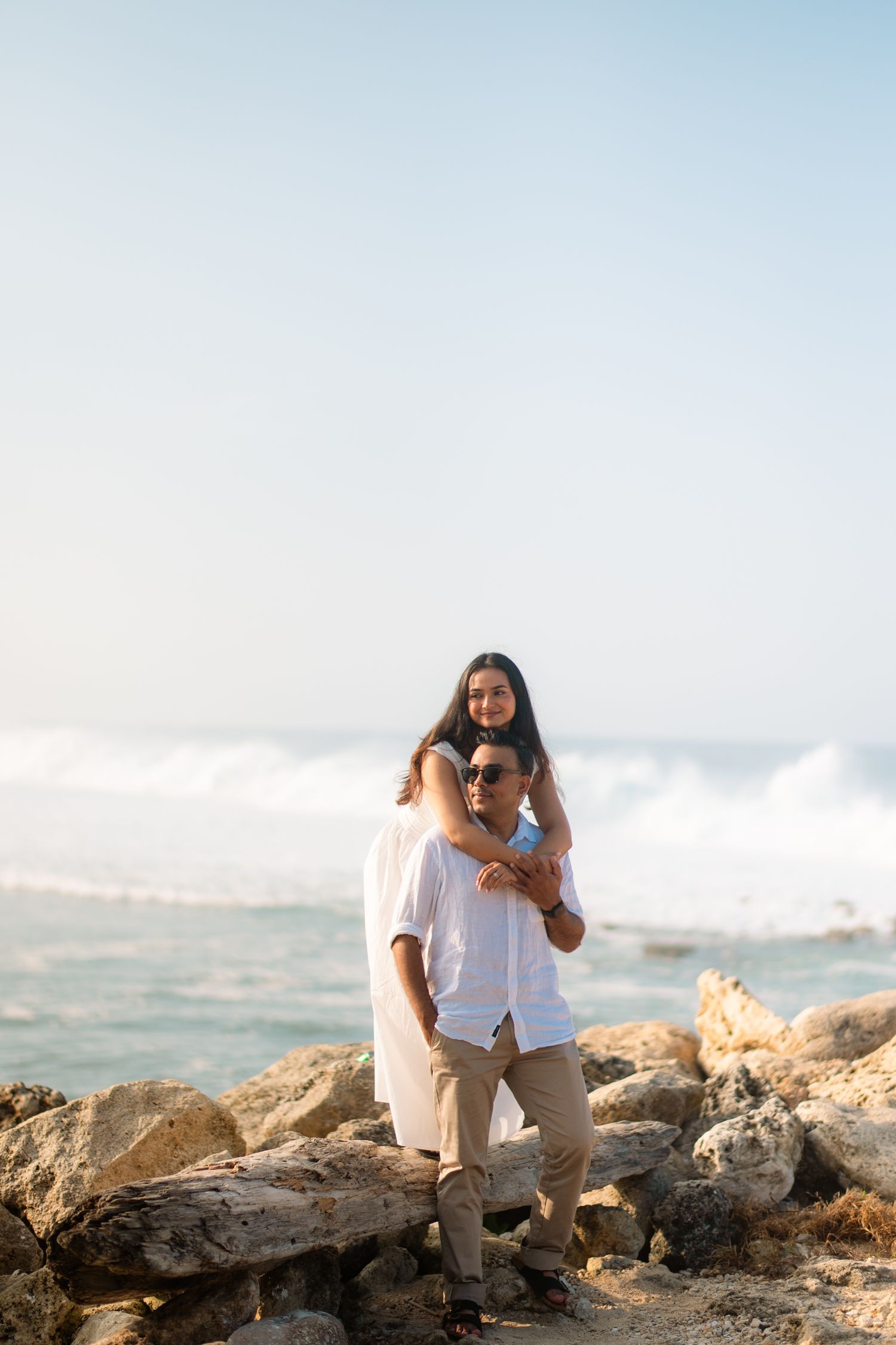 Couple embracing on rocky beach with ocean waves, woman in white dress hugging man from behind in sunny coastal setting.