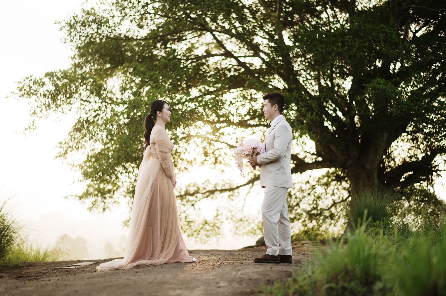 Couple sharing a romantic moment under a large tree on a misty hilltop during a dreamy outdoor shoot.