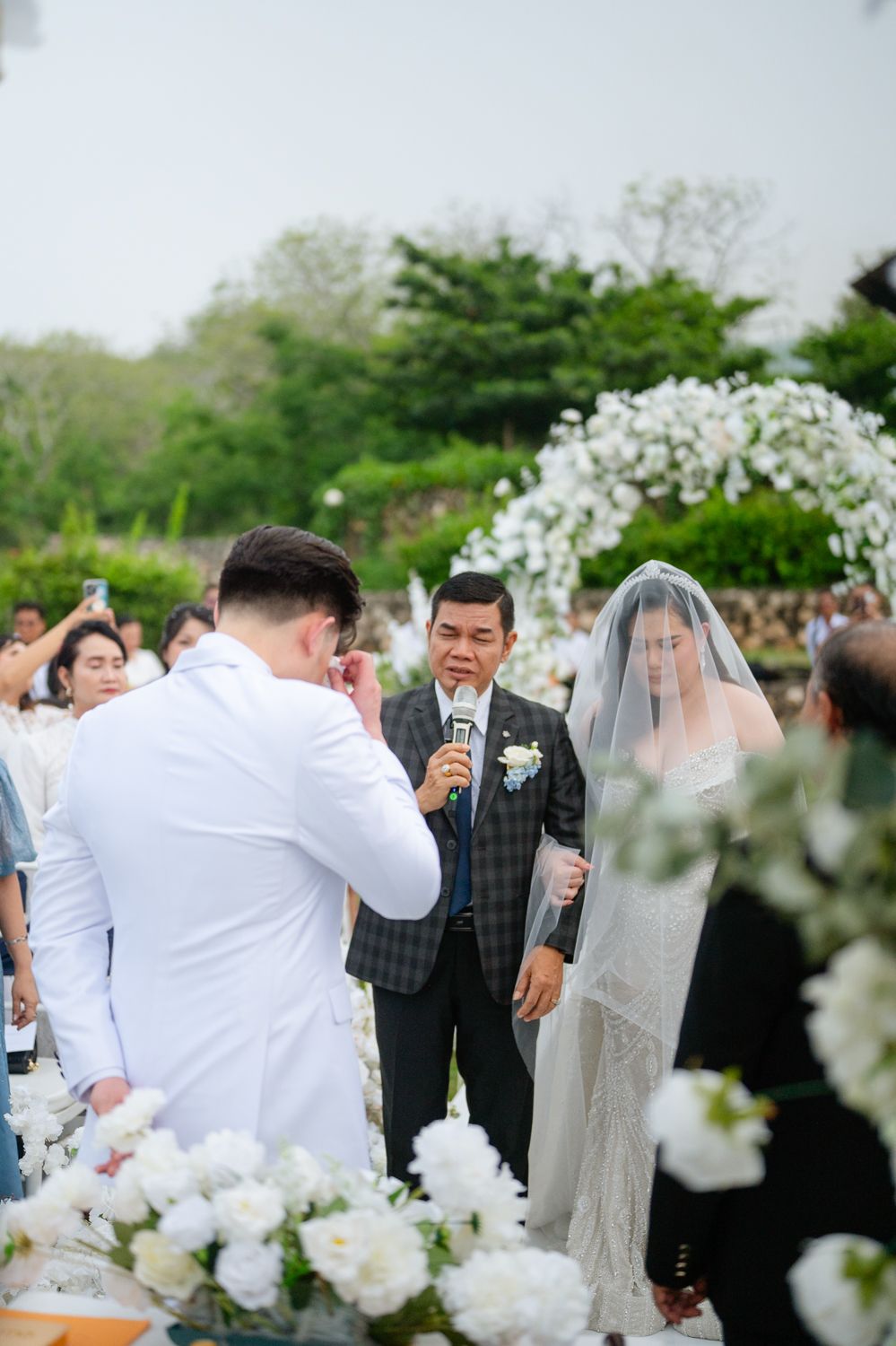 Outdoor wedding ceremony with bride in white veil, groom in white suit, and officiant in plaid suit amid white floral decorations.