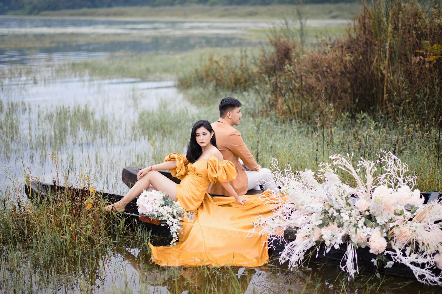 Couple in yellow dress sitting in a flower-decorated boat on a serene lake for a romantic photoshoot.