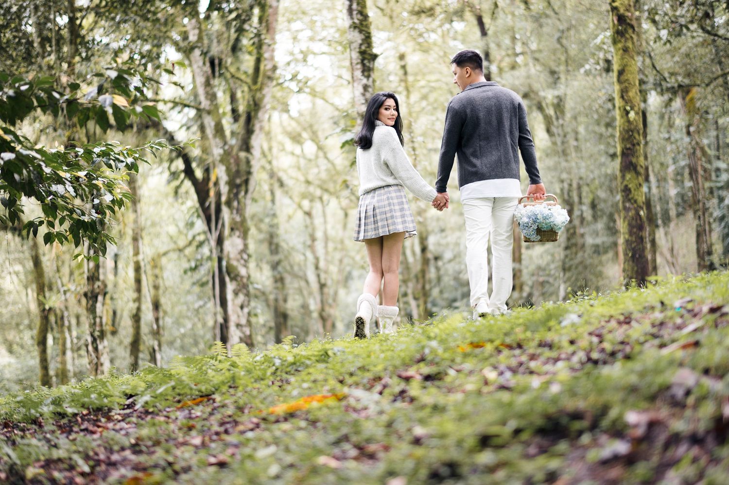 Couple walking hand in hand through a lush green forest path, woman in plaid skirt and white boots, man carrying flowers.